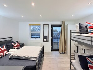 A compact hotel room with a bunk bed, a single bed, a window with a view of buildings, a radiator, and a door with a curtain. - Flat in Bethnal Green (London)