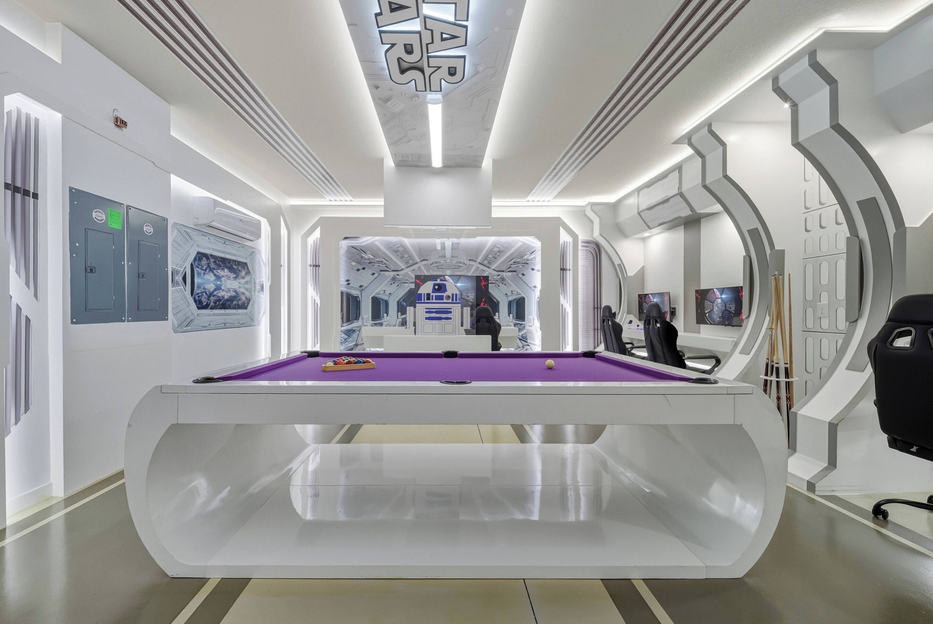 Ein moderner Innenbereich mit Schwimmbad, einem violetten Billardtisch, weißen Wänden und Star Wars Dekoration.