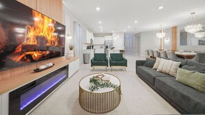 Uma sala de estar moderna com uma grande televisão de tela plana, uma mesa de centro redonda, um sofá cinza e uma área de jantar ao fundo.