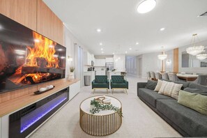 Ein modernes Wohnzimmer mit einem großen Flachbildfernseher, einem runden Couchtisch, einem grauen Sofa und einem Essbereich im Hintergrund.