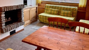 Een knus ingerichte kamer met een stenen openhaard, een groene geruite bank, een houten tafel en uitzicht op een vallei door een groot raam.