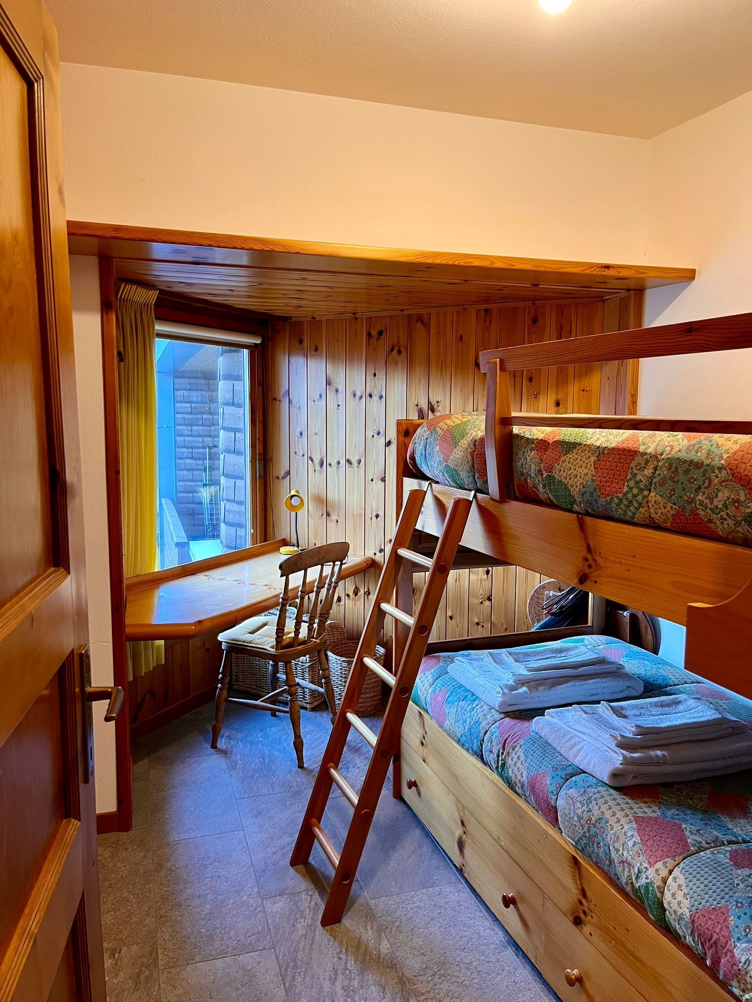 A room with a bunk bed, a desk with a chair, a ladder, and a window.