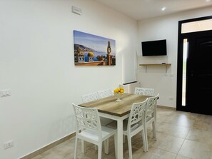 A dining area with a wooden table and white chairs, a painting of a coastal scene, a TV on the wall, and a black door. - As at home (Sicilia)