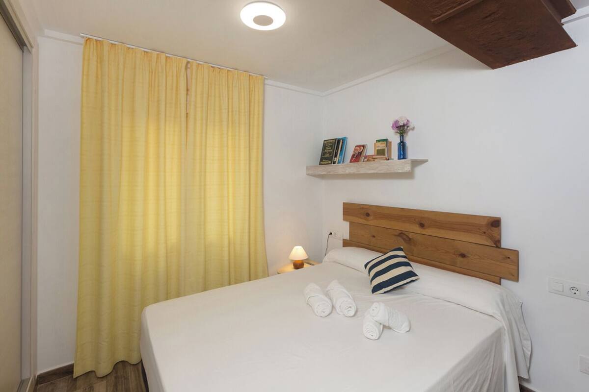 Una cama bien tendida con cabecera de madera, una lámpara de noche y una estantería con libros y un jarrón.