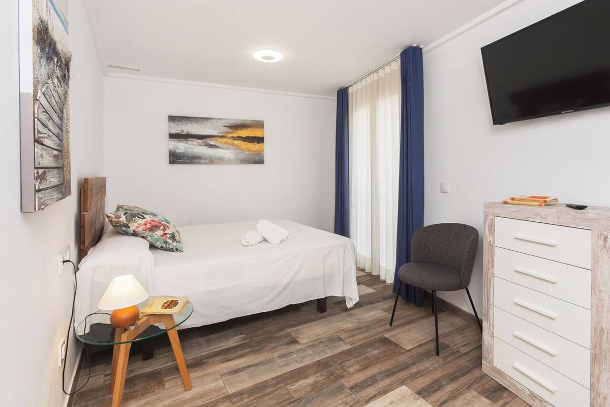 Habitación de hotel con cama, escritorio con cajones, silla, lámpara, un cuadro en la pared y televisor.