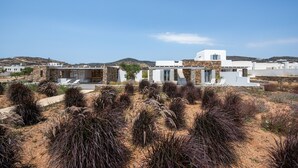 A modern white building with a stone facade, surrounded by dry vegetation and hills in the background. - Farous Seaside Sunset Villa II (Antiparos)