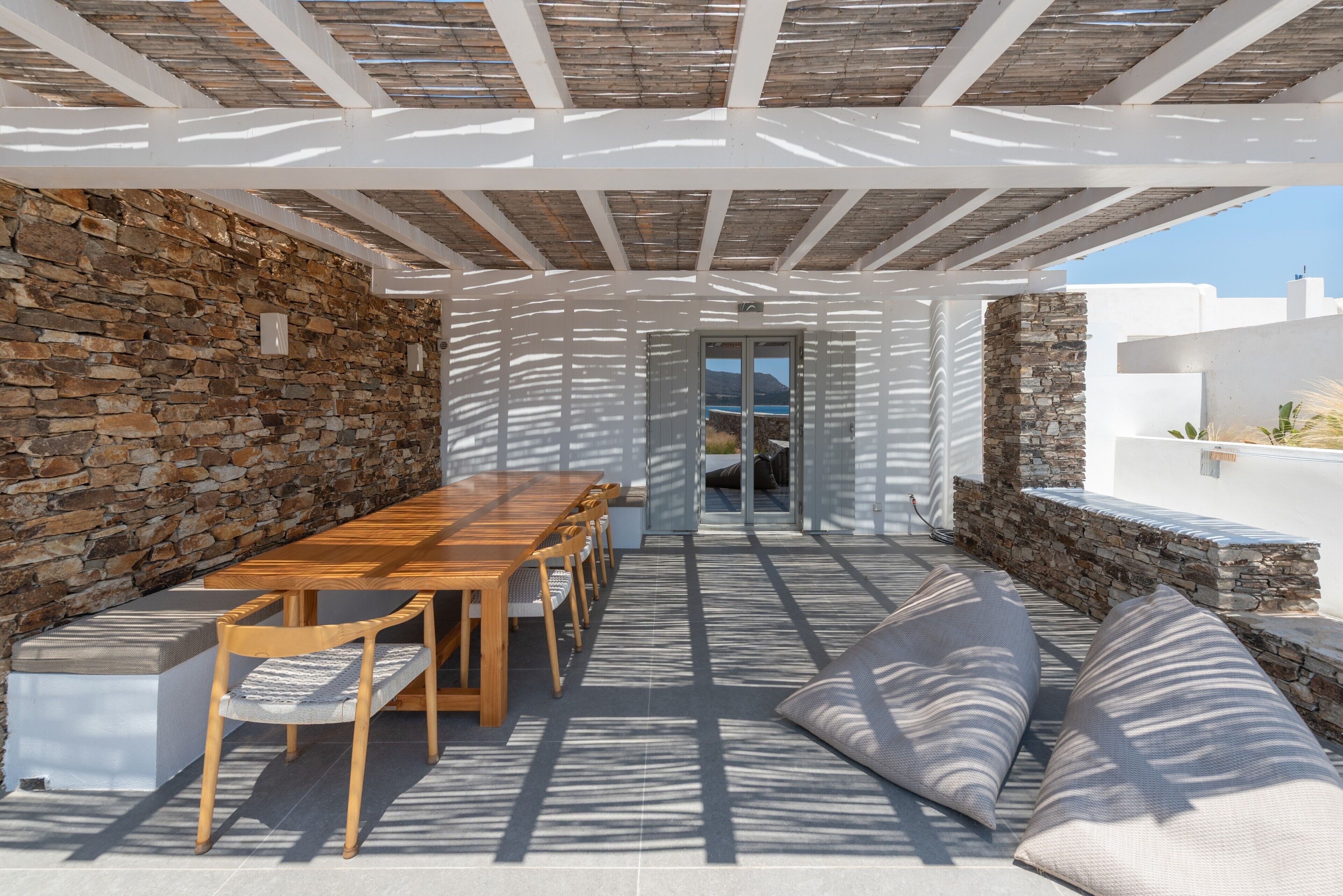 Een terras buiten met een houten tafel en stoelen, stenen muren en uitzicht op zee.