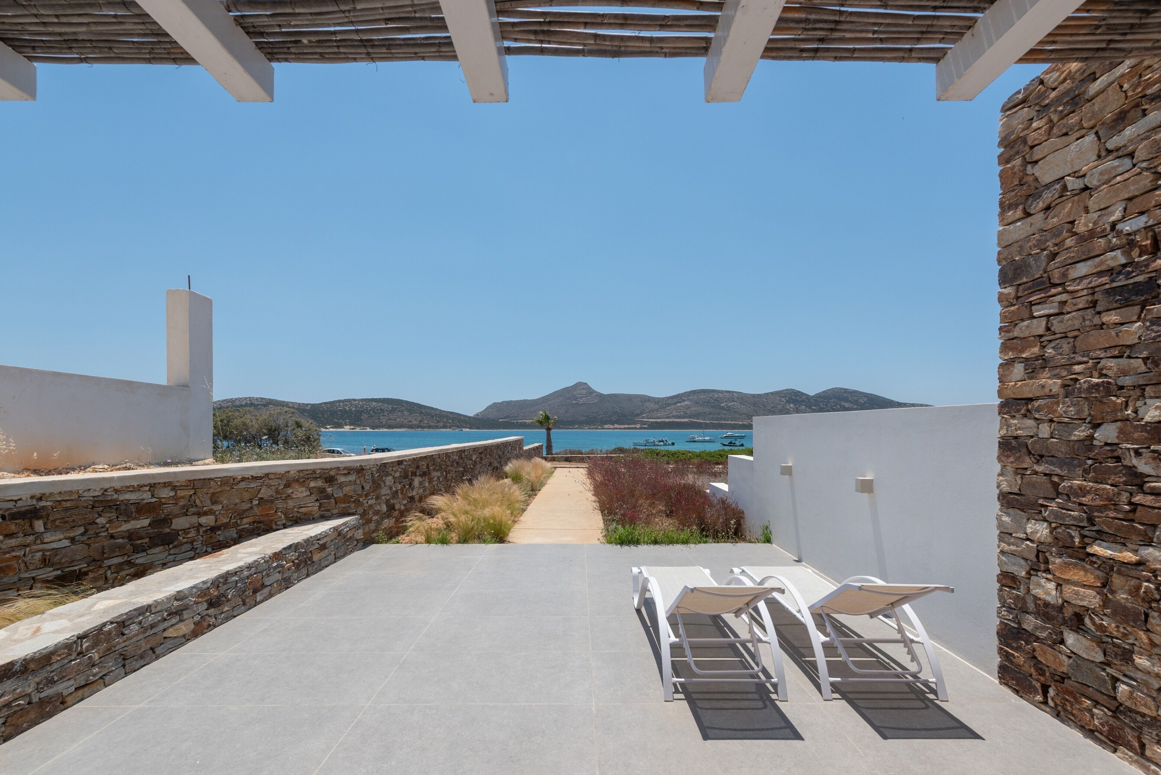 Une terrasse avec des murs en pierre, un sol en béton et deux fauteuils blancs donnant sur un paysage magnifique, avec la mer et les montagnes en toile de fond.