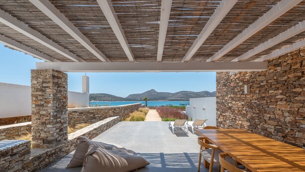 A terrace with stone walls, a wooden table, and chairs, overlooking a coastal view. - Farous Seaside Sunset Villa II (Antiparos)