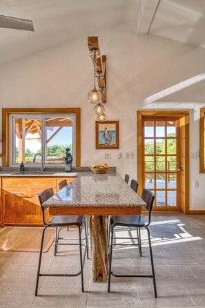 A dining area with a large table, chairs, a wooden island, and a view through large windows.