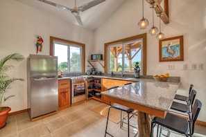 A kitchen with a stainless steel refrigerator, wooden cabinets, a granite countertop, a large island with bar stools, a ceiling fan, and a window with a view of greenery.