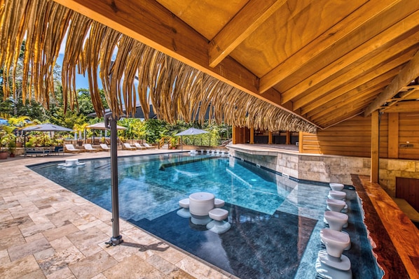 A swimming pool with a wooden deck, lounge chairs, and a thatched roof structure.