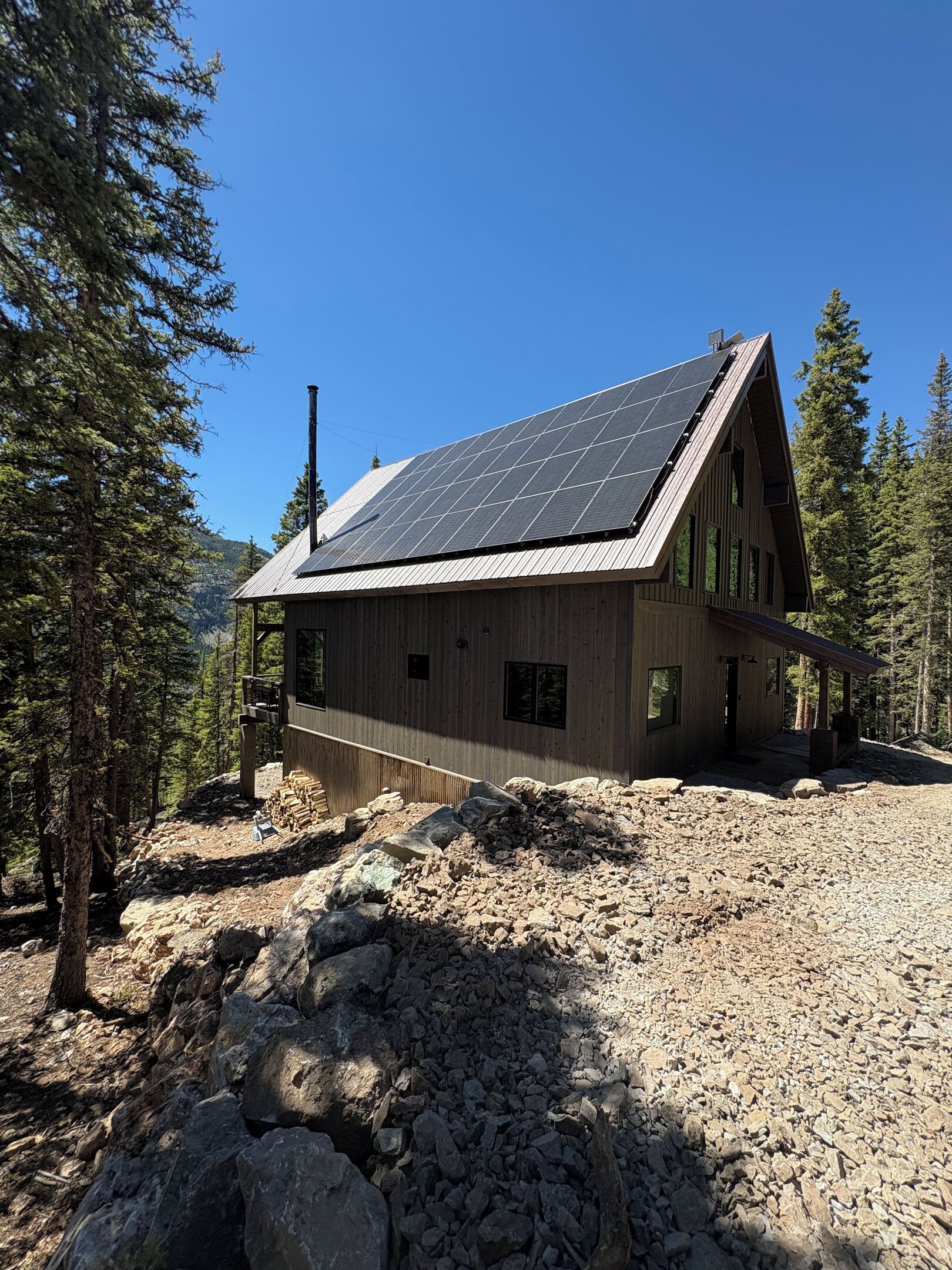 Une cabane en bois avec un toit en pente, des panneaux solaires et un terrain rocheux parsemé d’arbres.