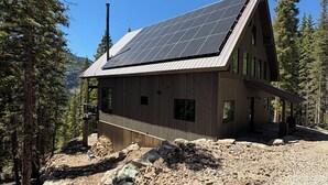 Une cabane en bois avec un toit en pente, des panneaux solaires et un terrain rocheux parsemé d’arbres.