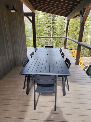 Eine überdachte Terrasse im Freien mit einem langen Tisch und Stühlen, umgeben von Holzgeländern und Blick auf Bäume.