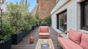 Una terraza en la azotea con una chimenea de ladrillo, un suelo de madera, dos sillones con cojines rosados, una mesita y plantas en macetas.