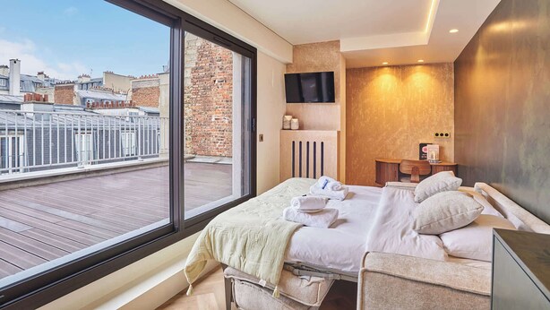 Una habitación de hotel moderna con un balcón amplio, una cama con sábanas blancas, un televisor y una mesita de noche de madera.