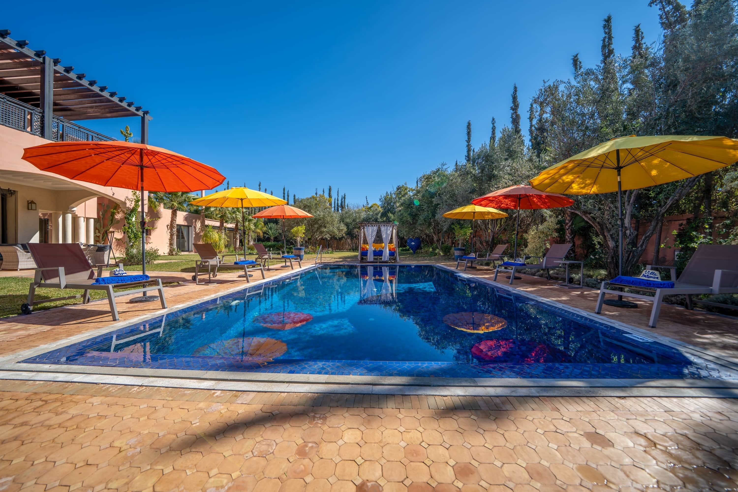 A swimming pool with lounge chairs and umbrellas, surrounded by trees and a building.