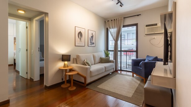 A modern living room with a sofa, a coffee table, and a TV stand. There is a window with curtains, a door leading to another room, and a plant on the windowsill. - Near Rodrigo de Freitas Lagoon Pc308 (Rio de Janeiro)
