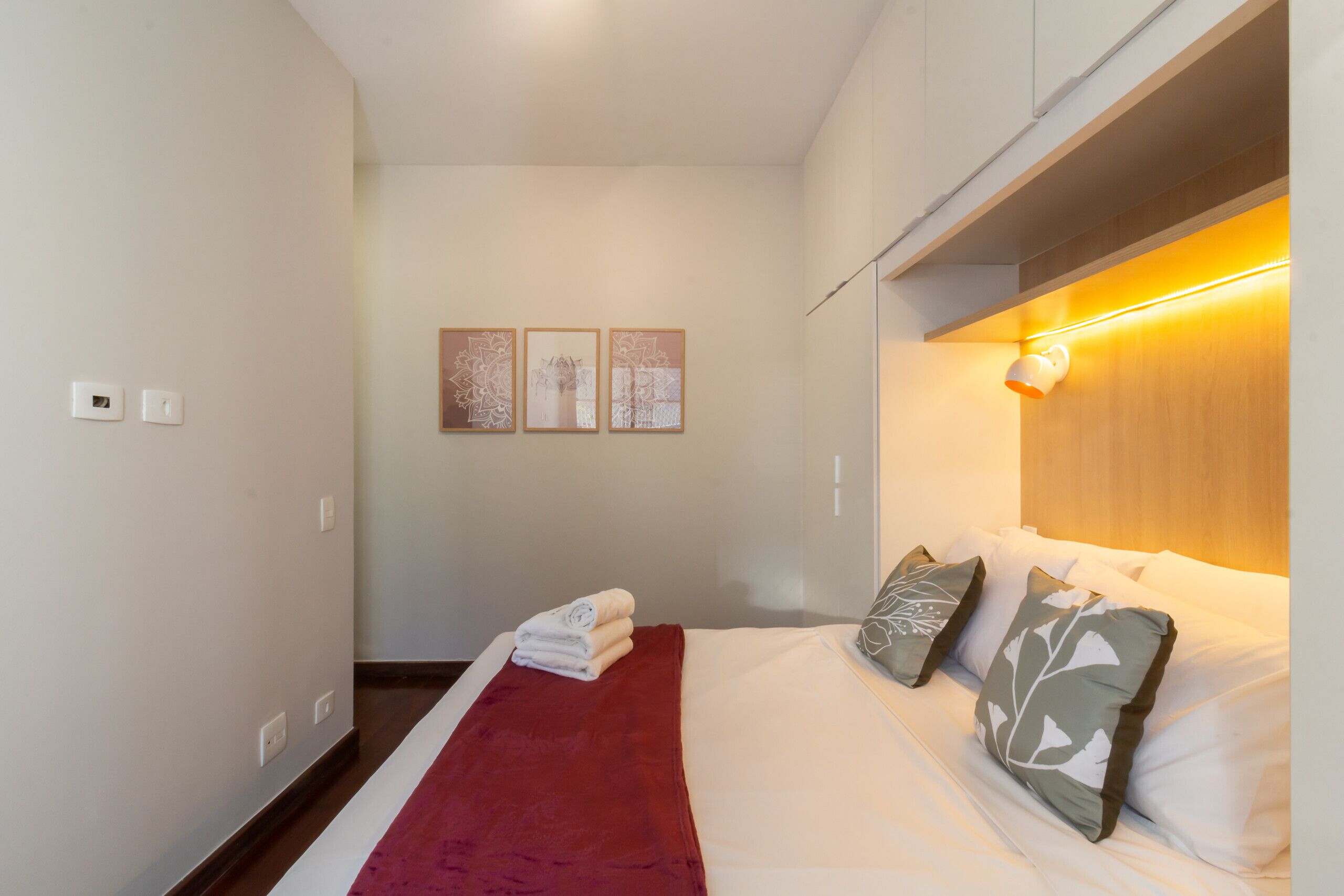 A hotel room with a bed, a red throw blanket, and three wall-mounted artworks.