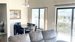 Ein Wohnzimmer mit grauem Sofa, einem Esstisch mit Stühlen, einer Kristallleuchtre und einem großen Fenster mit Aussicht.