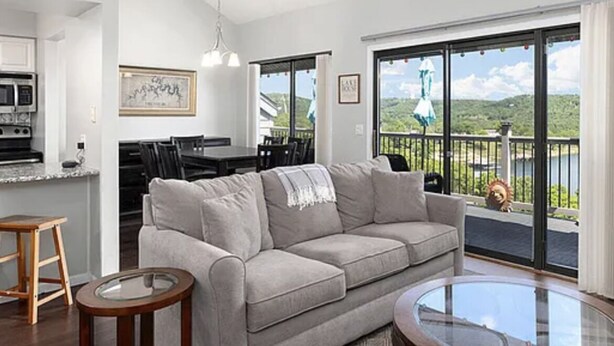 Ein großzügiger Wohnbereich mit grauem Sofa, einem Glastisch und einem Esstisch mit Stühlen. Es gibt eine Küchenzeile mit Mikrowelle und Schränken, sowie einen Balkon mit Landschaftsblick.