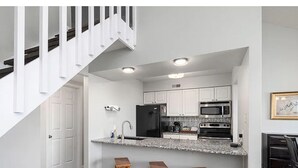 Eine moderne Küche mit weißen Schränken, einer Granit Arbeitsplatte und einem schwarzen Kühlschrank. Drei hölzerne Barhocker stehen an der Theke.