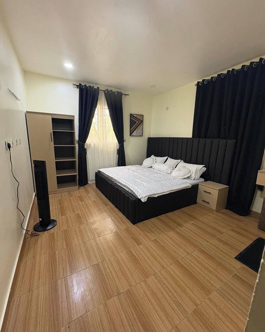 Un dormitorio con cama, mesita de noche, armario y ventilador de pie.