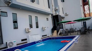 An indoor swimming pool with a blue tiled border, air conditioning units, and outdoor seating with umbrellas. - CORA HOMES (Jos)