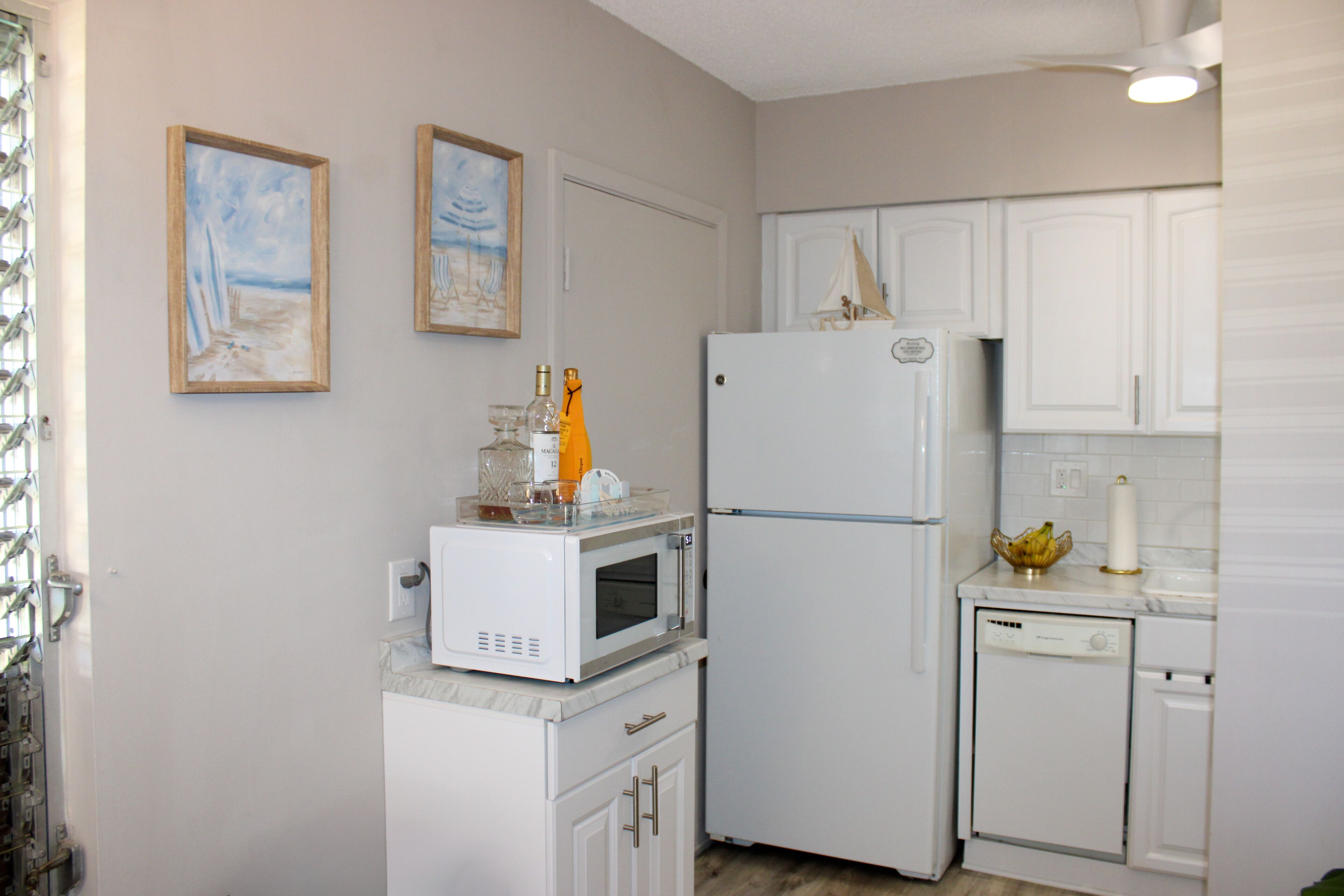 A compact kitchen with white cabinets, a microwave, a refrigerator, and a dishwasher. There are two framed paintings on the wall.