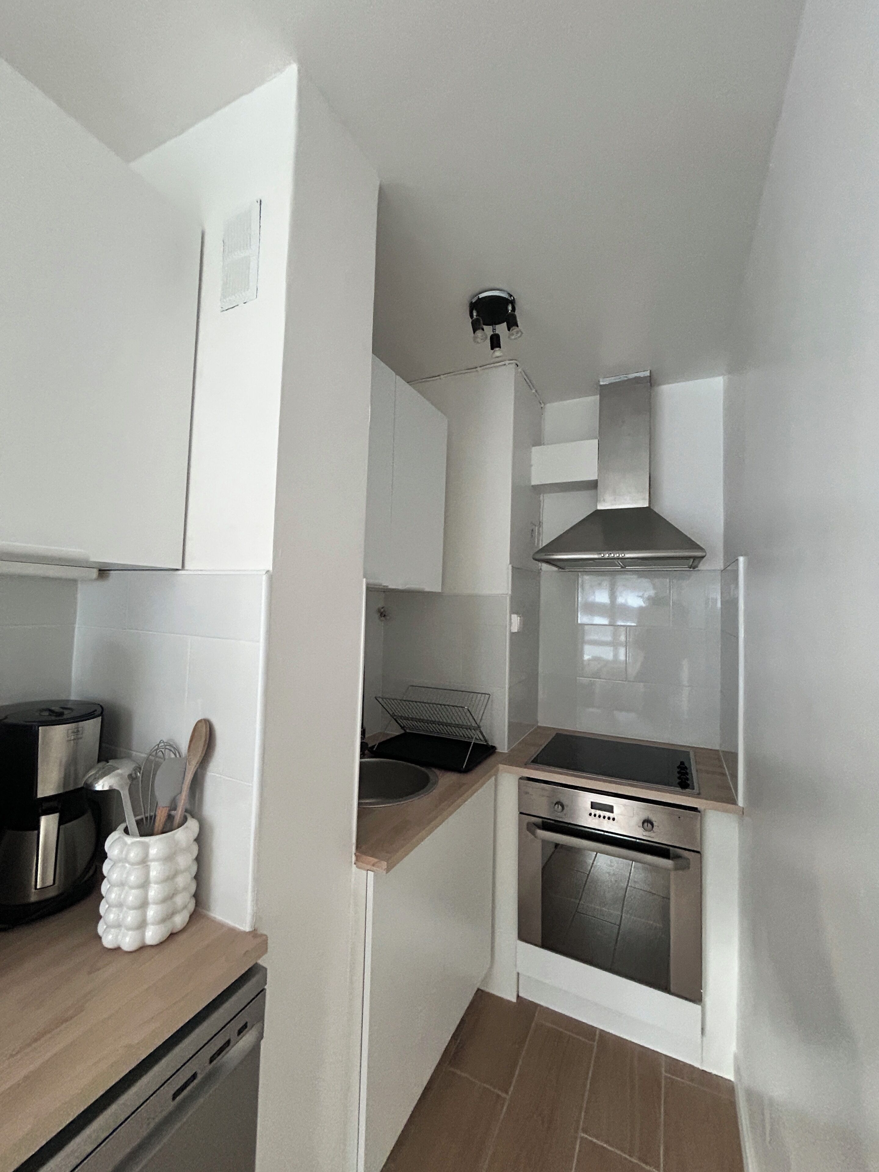 A compact kitchen with white cabinets, a stainless steel range hood, a built-in oven, and a coffee maker.