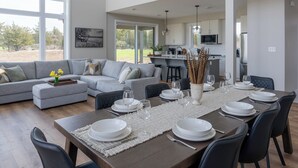 Un área de comedor moderna con una mesa larga preparada para seis personas, un sofá gris y una cocina al fondo.