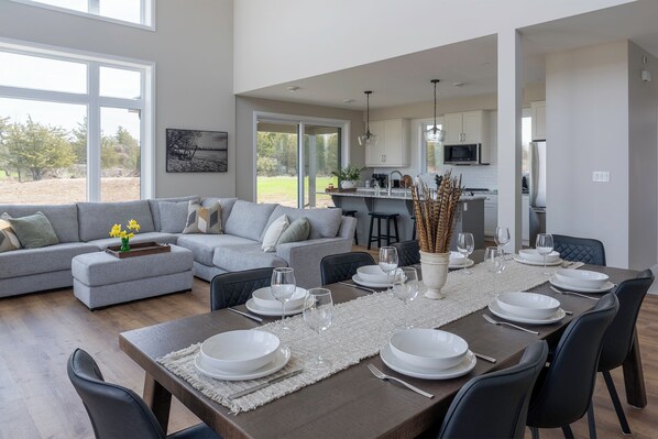A modern dining area with a long table set for six, a grey sofa, and a kitchen in the background.