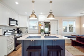 A modern kitchen with white cabinets, stainless steel appliances, and a central island with stools. - Pickled Pelican: Bright and Airy Oceanfront Home (Oak Island)