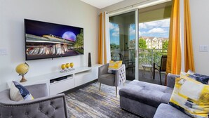 Ein modernes Wohnzimmer mit Flachbildfernseher, einer Couch, einem Sessel, einem kleinen Tisch und einem Balkon mit gelben Vorhängen.
