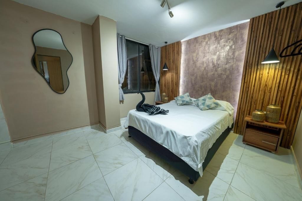 Une chambre à coucher avec un lit, un miroir, une fenêtre avec des rideaux, une table de chevet et une lampe.