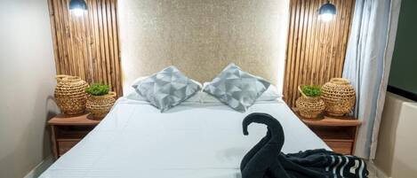 A neatly made bed with two pillows, a black swan-shaped towel, and two wicker baskets with plants on either side.