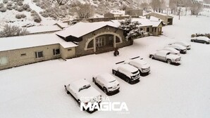 Un estacionamiento cubierto de nieve con varios coches, un edificio con techo inclinado y montañas al fondo.