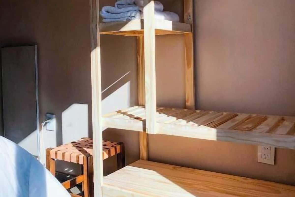Una cama litera con listones de madera, una mesita pequeña y un perchero en una habitación con pared lisa y una puerta.