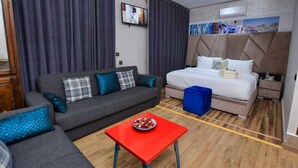 A modern hotel room with a grey sofa, a red table with a bowl of fruit, a large bed with a headboard, and a TV mounted on the wall. - Hotel Dar Annasr (Chefchaouen)