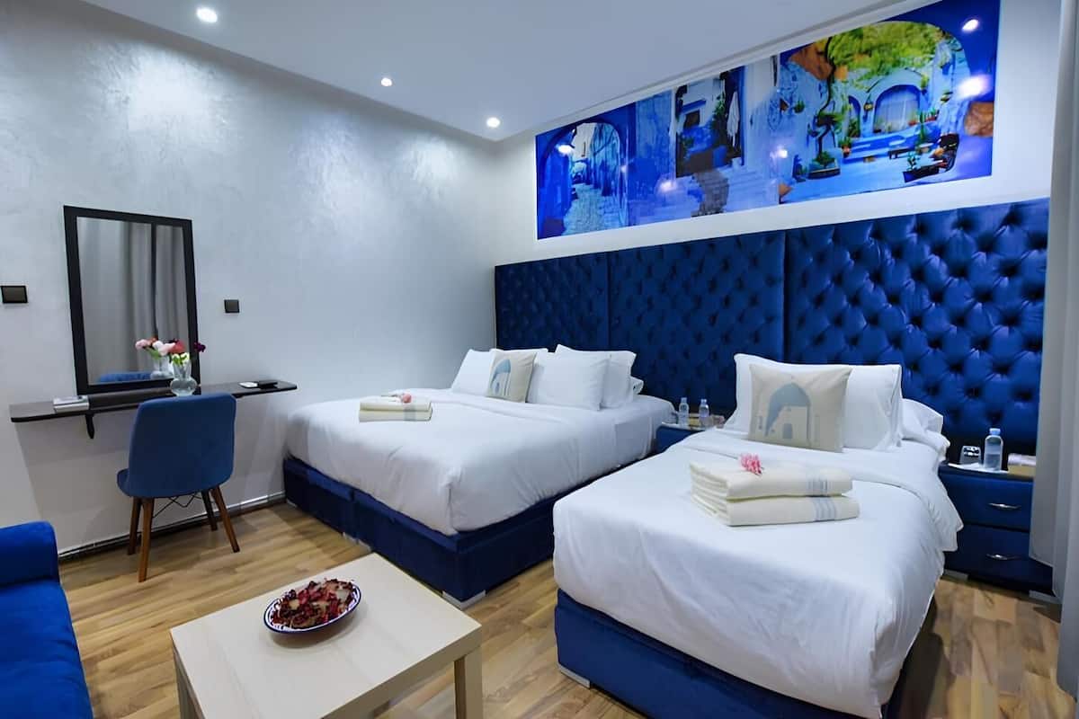 Une chambre d’hôtel avec deux lits, un bureau, une chaise et une petite table sur laquelle se trouve un saladier rempli de fruits.