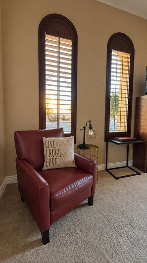 A room with a leather armchair, a small table with a lamp, and two arched windows with blinds.