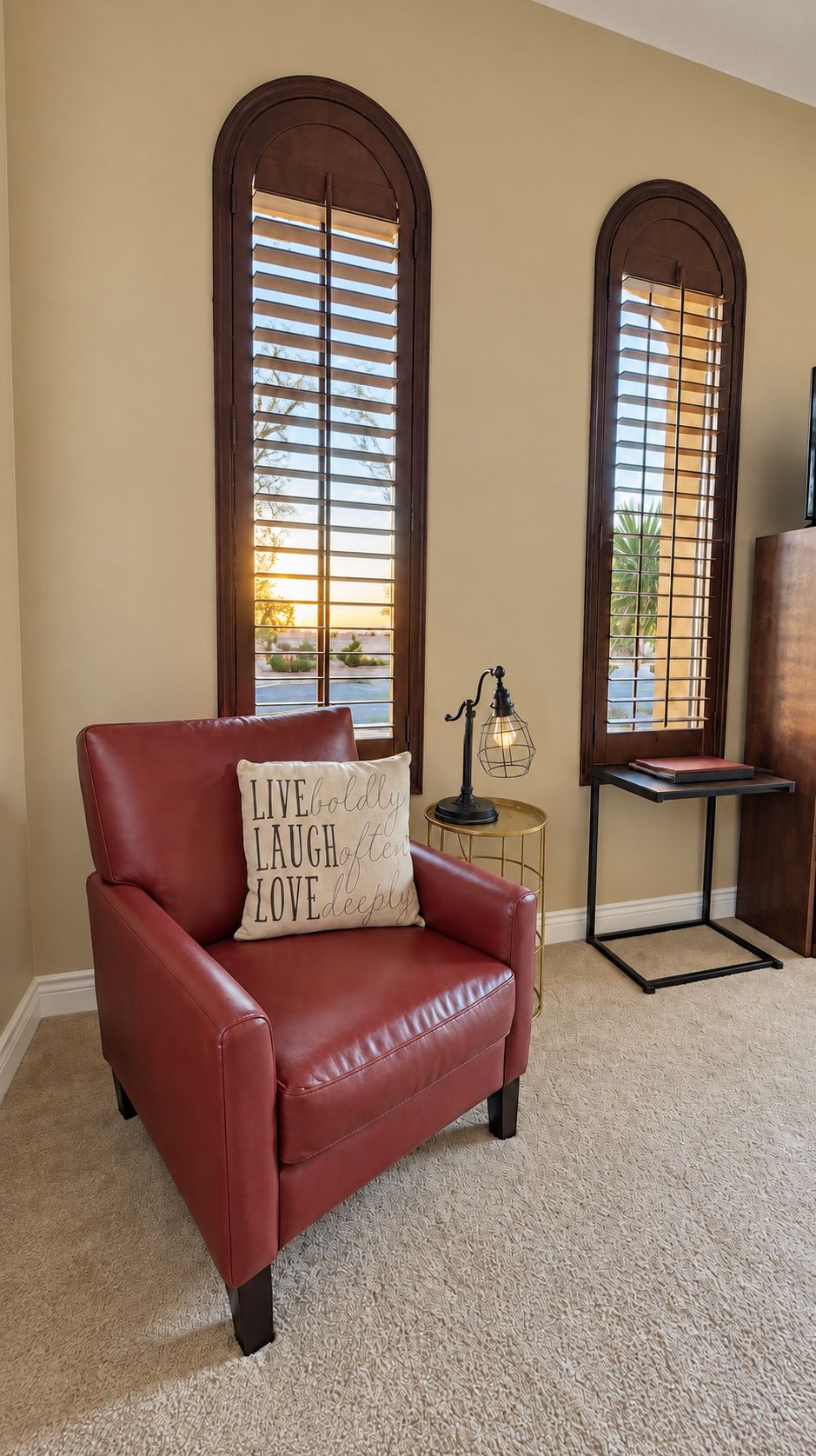 A room with a leather armchair, a small table with a lamp, and two arched windows with blinds.