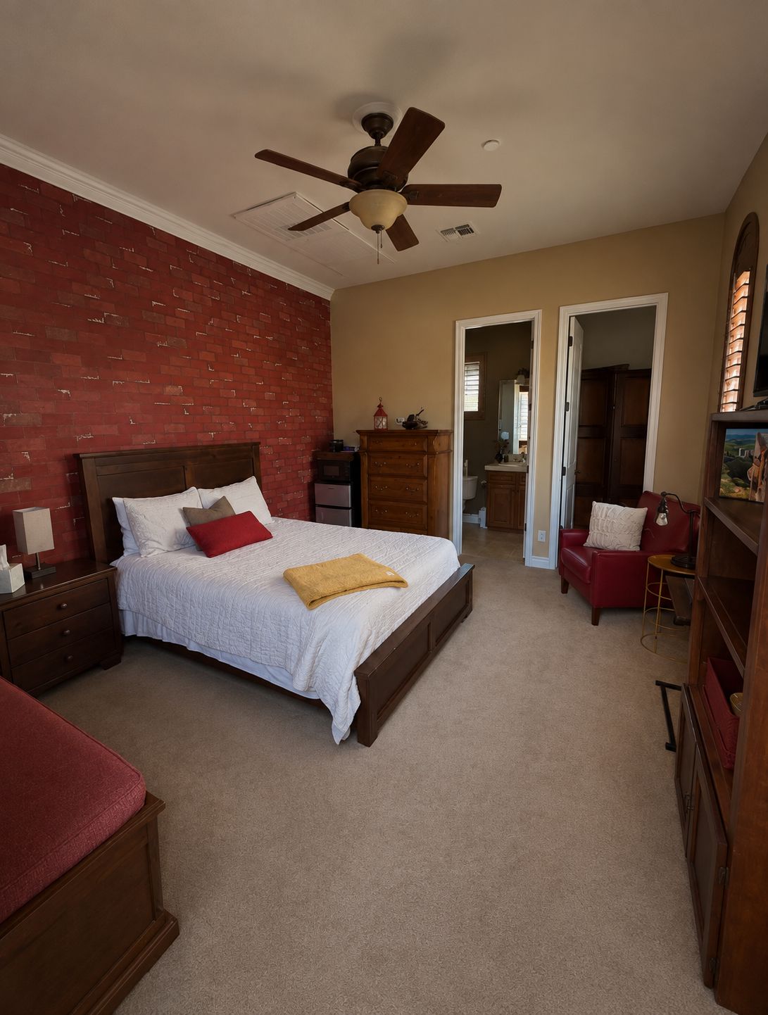 Une chambre à coucher avec un grand lit, un ventilateur de plafond, une commode, un fauteuil rouge et une bibliothèque.
