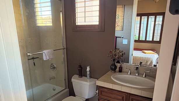 Une salle de bain avec une douche à parois de verre, des toilettes, un lavabo avec un miroir et une fenêtre avec des stores.