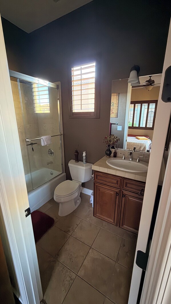 Een badkamer met een douchecabine van glas, een toilet, een wastafel met spiegel en een raam met jaloezieën.