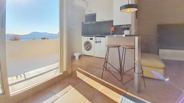 A compact kitchen with a washing machine, a small dining area with stools, and a view of the sea through a large window. - Breathtaking panoramic sea view - Exceptional 2P at Le Grand Duc (Mandelieu-la-Napoule)