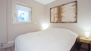 A bedroom with a large bed, a window, a bedside table with a lamp, and a painting on the wall. - Breathtaking panoramic sea view - Exceptional 2P at Le Grand Duc (Mandelieu-la-Napoule)