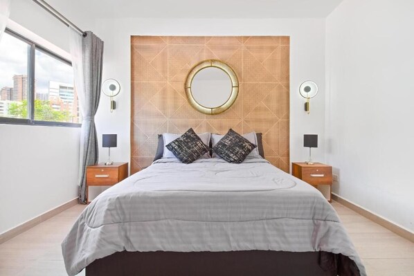 Camera da letto moderna con un letto grande, due comodini, uno specchio rotondo e vista sulla città attraverso una finestra.