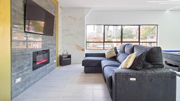 A modern living room with a grey sofa, a flat-screen TV mounted on a textured wall, and a large window offering a city view. - 2BR w Pool Table 1 Block from Lleras (Medellín)